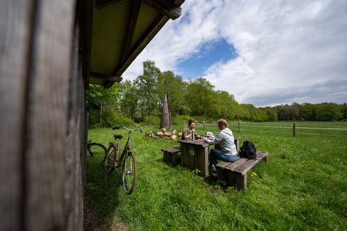 Zwei Radfahrer trinken auf einem malerischen Rastplatz am Ochsenweg bei Neumünster einen Kaffee, im Hintergrund ist eine Ochsen-Hörner-Skulptur zu sehen.