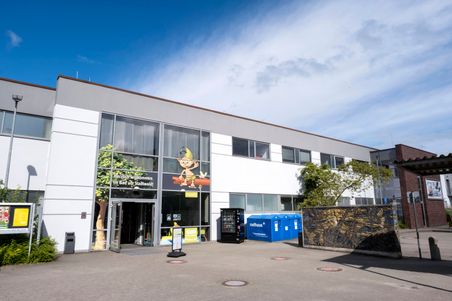 Das Bild zeigt den Eingang des 'Bad am Stadtwald', ein modernes Gebäude mit Glasfront und einer Cartoon-Figur. Vor dem Eingang stehen Müllcontainer.