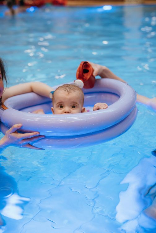 Ein Baby sitzt in einem lilafarbenen Schwimmring im Pool, während eine erwachsene Person es hält. Das Wasser ist klar und blau.
