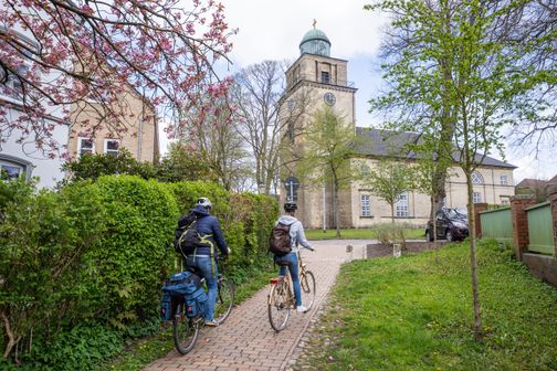 Zwei Radfahrer fahren auf einem gepflasterten Weg zur Vicelinkirche in Neumünster. Blühende Bäume und grüne Büsche säumen den Weg.