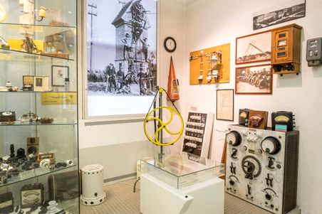 Ein Raum im Elektromuseum Rendsburg zeigt historische elektrische Geräte, Fotos und Schautafeln. Ein gelbes Rad und Messinstrumente sind zu sehen.