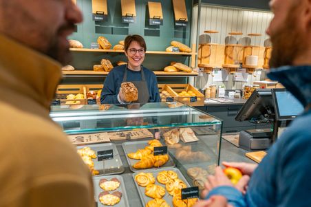 Eine freundliche Bäckereiverkäuferin präsentiert ein Brot hinter der Theke. Verschiedene Backwaren sind im Hintergrund zu sehen.