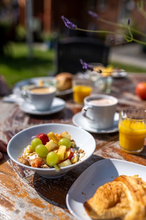 Ein einladender Frühstückstisch im Freien mit Müsli, Croissant, Kaffee und Orangensaft. Lavendelzweige ragen ins Bild.