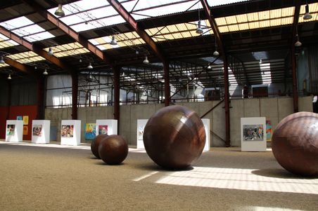 Große Metallkugeln und Gemälde in einer lichtdurchfluteten Ausstellungshalle mit Oberlichtern und industrieller Architektur.