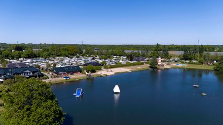 Luftaufnahme des Familien-Campingplatzes Padenstedt mit Wohnwagen, einem See, Strand und Spielplatz. Ein Leuchtturm und Wasserspielgeräte sind sichtbar.