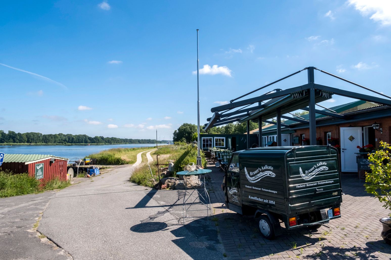 Brauers Aalkate liegt idyllisch am Flussufer. Ein kleiner Lieferwagen steht vor dem Restaurant, das von grüner Natur umgeben ist.