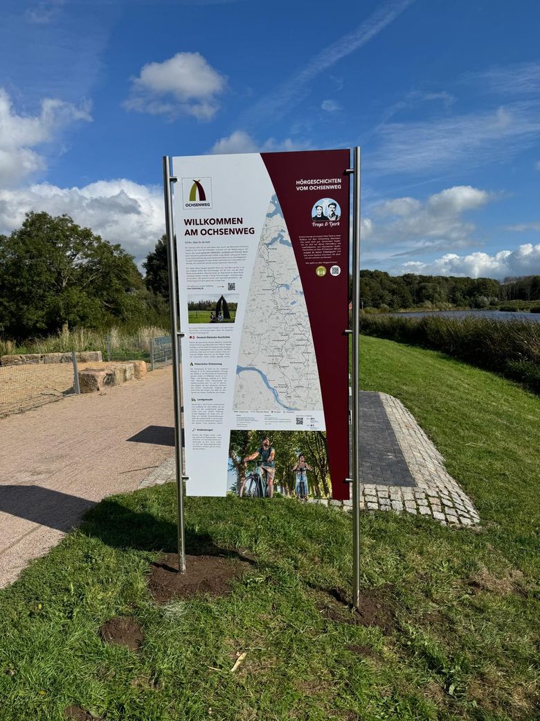 Ein Informationsschild am Ochsenweg mit Karte und Text, umgeben von grüner Landschaft und blauem Himmel. Es begrüßt Besucher und bietet Informationen zur Route.
