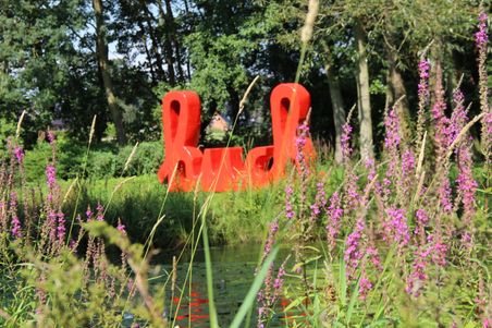 Ein roter Schriftzug steht im Gerisch-Park hinter lila blühenden Blumen und Gräsern, umgeben von Bäumen.