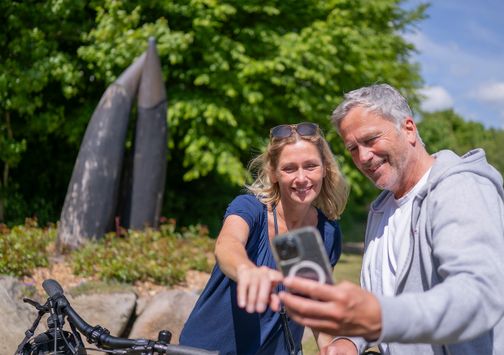 Ein Mann und eine Frau machen ein Selfie vor dem Hörnerplatz im Grünen. Beide lächeln und wirken entspannt. Ein Fahrrad ist im Vordergrund zu sehen.
