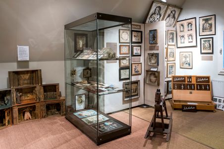 Ein Ausstellungsraum im Heimatmuseum Hohenwestedt zeigt historische Exponate in Vitrinen und an den Wänden. Ein Schaukelpferd und ein Schokoladenspiel sind zu sehen.