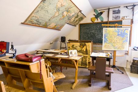Ein nostalgisches Klassenzimmer mit alten Karten, einer Tafel voller Kritzeleien und klassischen Schulbänken. Ein Mikroskop und Lehrmaterialien sind sichtbar.