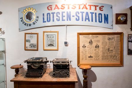 Zwei antike Schreibmaschinen auf einem Holztisch, umgeben von historischen Schildern und gerahmten Dokumenten in einem Museum.