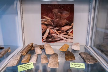 Flintbeile in einer Vitrine ausgestellt, mit Schildern "Gebrauchsfertige Flintbeile" und "Nachgeschliffene Flintbeile". Im Hintergrund ein Bild von Keramik.
