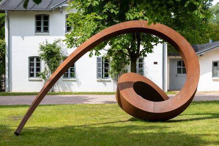 Rostfarbene Spiral-Skulptur auf einer Wiese vor einem weißen Gebäude mit Fenstern. Die Skulptur ist groß und geschwungen, umgeben von Bäumen.