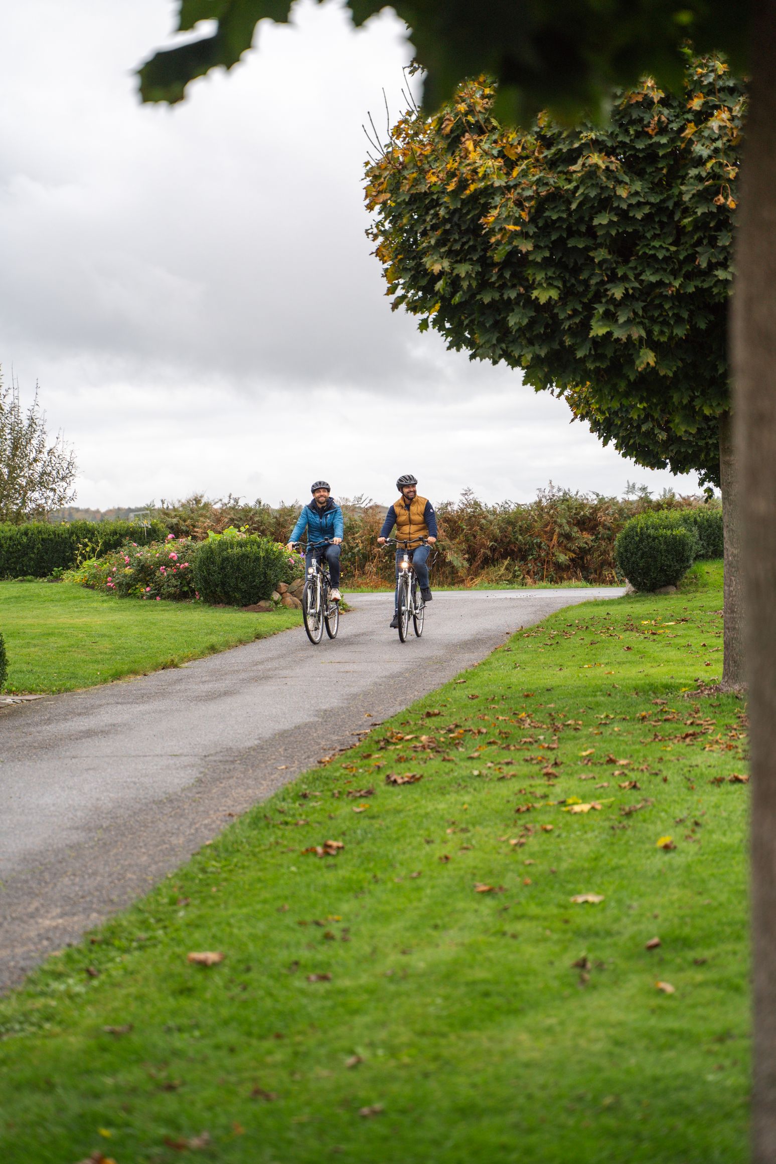 Zwei Radfahrer auf einem Weg, umgeben von grüner Landschaft und Bäumen. Der Himmel ist bewölkt, und es ist Herbst.