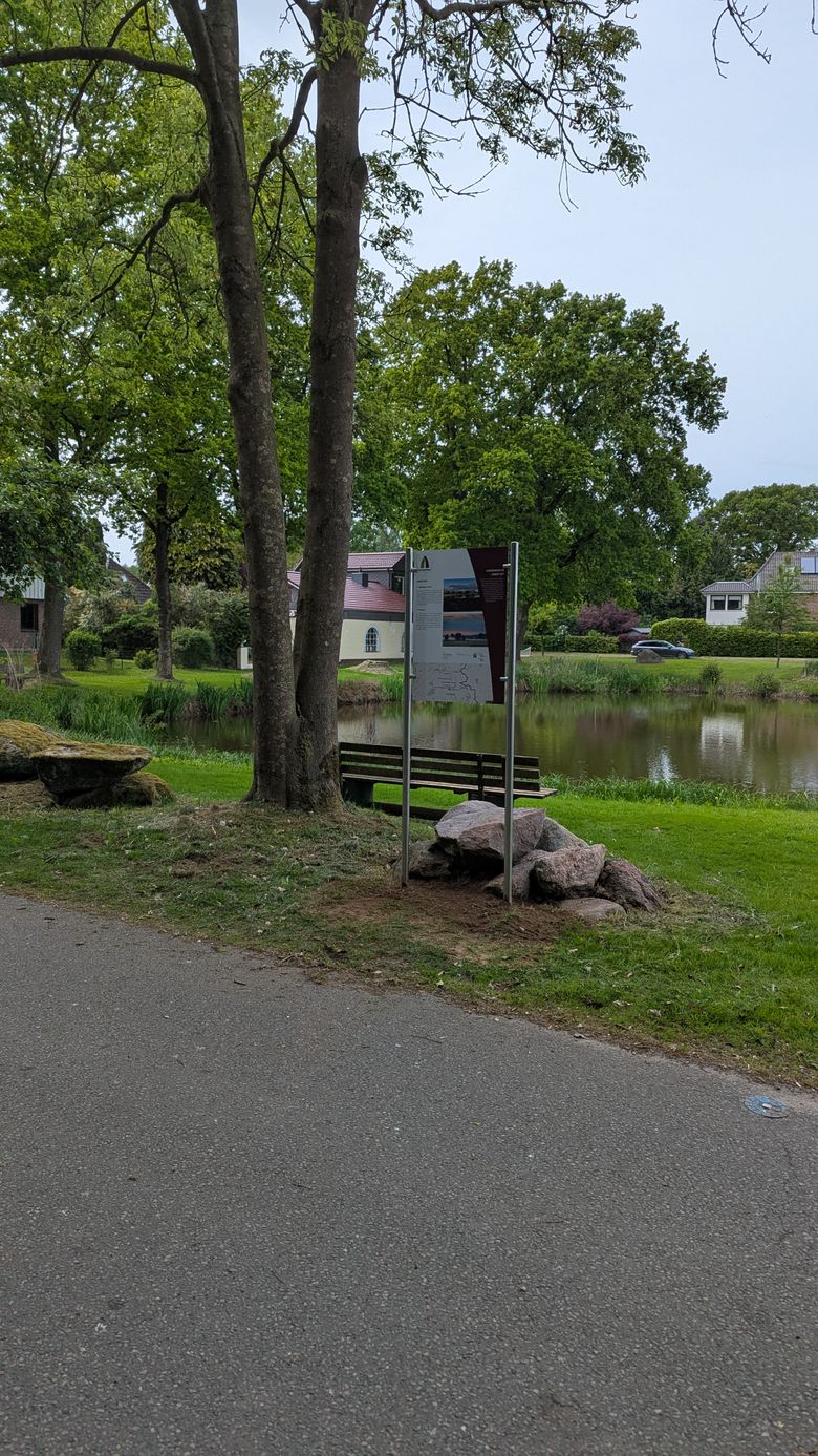 Ein ruhiger Weg führt an einem Teich vorbei, umgeben von grünen Bäumen und Häusern. Eine Infotafel steht neben einem Baum.