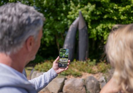 Ein Mann hält ein Smartphone und fotografiert die Hörner im Freien. Die Skulptur besteht aus zwei großen, spitzen Elementen vor grünem Hintergrund.