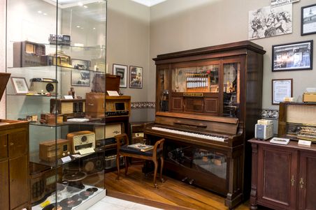 Ein Raum im Elektromuseum Rendsburg zeigt antike Radios in einer Vitrine und ein mechanisches Klavier. An den Wänden hängen historische Bilder.