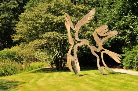 Eine Metallskulptur zeigt zwei stilisierte Vögel, die sich zu küssen scheinen. Sie steht auf einer grünen Wiese vor dichtem Laubwerk.