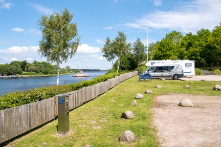 Ein Wohnmobil steht auf einem Stellplatz am Wasser. Im Hintergrund fährt ein Schiff vorbei. Der Platz ist von Bäumen und einem Holzzaun umgeben.