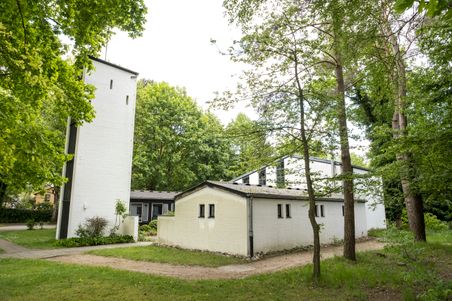 Die Kreuzkirche Schülp ist ein weißes Gebäude mit einem Turm, umgeben von Bäumen und Grünflächen. Der moderne Bau fügt sich harmonisch in die Natur ein.