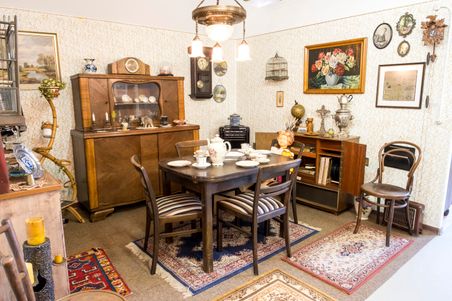 Ein nostalgisch eingerichtetes Wohnzimmer mit antiken Möbeln, Teppichen und Dekorationen. Ein Tisch ist gedeckt, umgeben von Stühlen und einem Schrank.