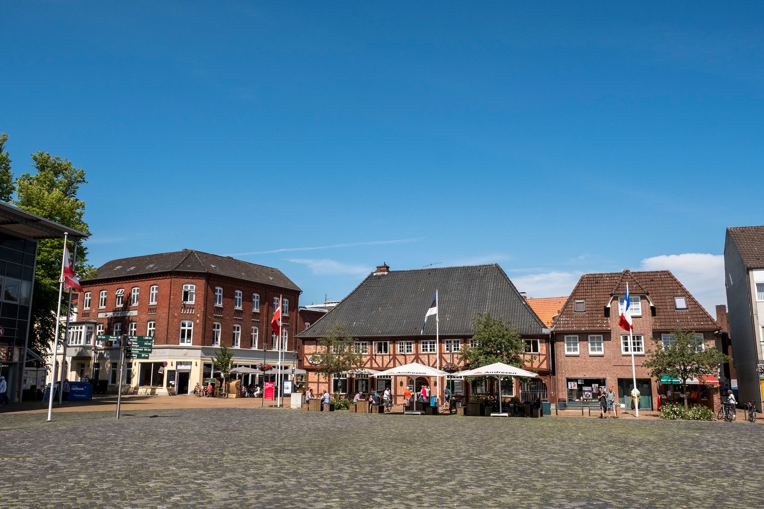 Der Schiffbrückenplatz in Rendsburg zeigt historische Gebäude und Flaggen. Menschen sitzen draußen bei sonnigem Wetter.