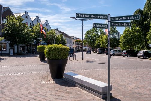 Ein Wegweiser in Nortorf zeigt in verschiedene Richtungen, darunter Bahnhof, Stadtpark und Polizeizentralstation. Im Hintergrund sind Gebäude und Autos sichtbar.