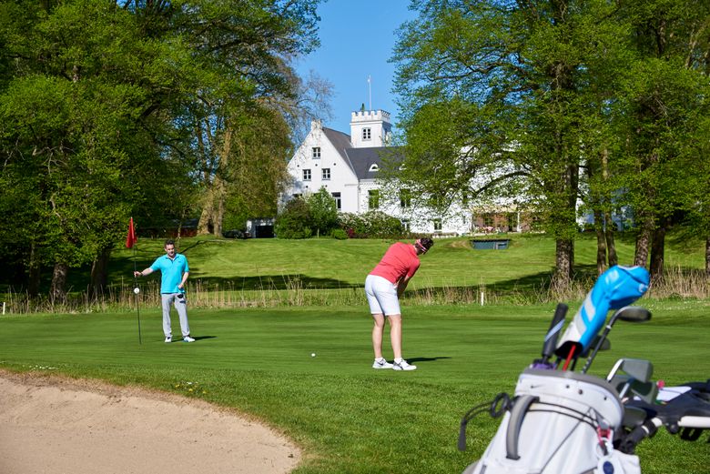 Zwei Golfer spielen auf einem Golfplatz vor dem Schloss Breitenburg, umgeben von Bäumen und blauem Himmel.