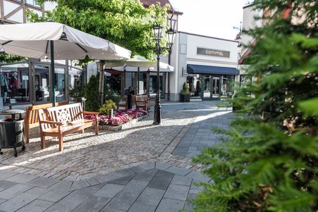 Ein gemütlicher Bereich im Neumünster Outlet-Center mit Bänken, Blumen und Geschäften. Sonnenschirme spenden Schatten.