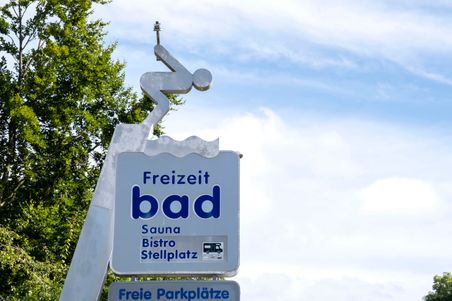 Ein Schild eines Freizeitbads mit einem Taucher-Piktogramm. Es bietet Sauna, Bistro und Stellplatz. Der Himmel ist blau mit einigen Wolken.