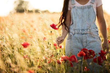 Eine Person in Jeanslatzhose steht in einem blühenden Mohnfeld und hält eine rote Mohnblume. Die Szene ist in warmes Sonnenlicht getaucht.
