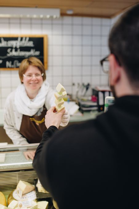 Eine freundliche Verkäuferin hinter einer Käsetheke reicht einem Kunden einen Spieß mit verschiedenen Käsestücken.