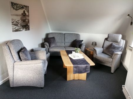 Gemütliches Wohnzimmer mit drei grauen Sesseln, Sofa, Holztisch und Wandbild „Glaube, Liebe, Hoffnung“.