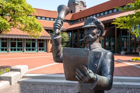 Bronzestatue eines Mannes mit Megafon und Papier vor einem Gebäude mit rotem Ziegeldach und Uhrturm in Hohenwestedt.