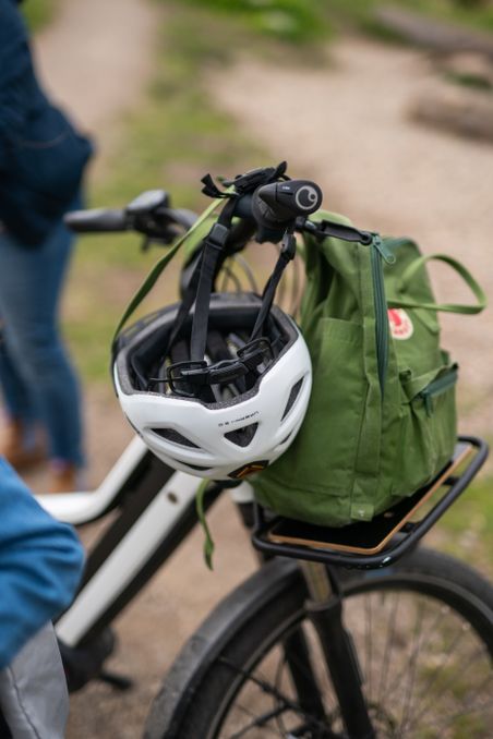 Ein Fahrrad mit einem weißen Helm und einem grünen Rucksack auf dem Gepäckträger steht auf einem Weg im Freien.