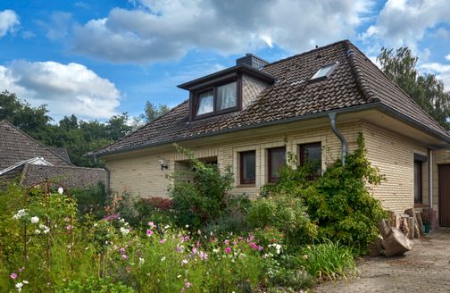 Haus mit Ziegeldach und blühendem Vorgarten unter blauem Himmel mit Wolken.