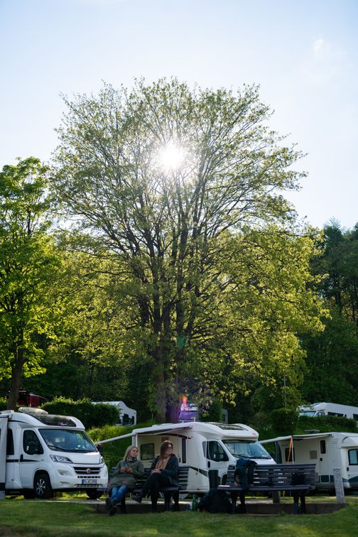 Zwei Personen entspannen auf einer Bank vor Wohnmobilen, umgeben von grünen Bäumen und strahlendem Sonnenschein.