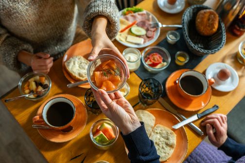 Ein reich gedeckter Frühstückstisch mit Kaffee, Brötchen, Aufschnitt und Obst. Zwei Personen teilen ein Glas mit Lachs.
