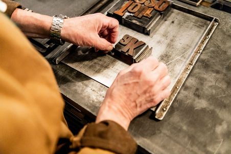 Hände ordnen Buchstaben in einer Druckform an, vermutlich in einem Druckmuseum. Die Szene zeigt traditionelle Drucktechniken.