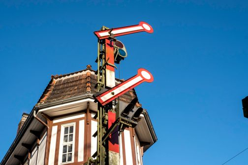 Ein historisches Stellwerk mit roten Signalarmen vor einem klaren blauen Himmel. Das Gebäude hat ein markantes Dach und Fachwerkelemente.