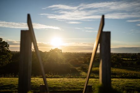 Ein Sonnenuntergang über einer weiten, grünen Landschaft. Im Vordergrund sind unscharfe Holzkonstruktionen zu sehen, die den Blick auf die Natur rahmen.