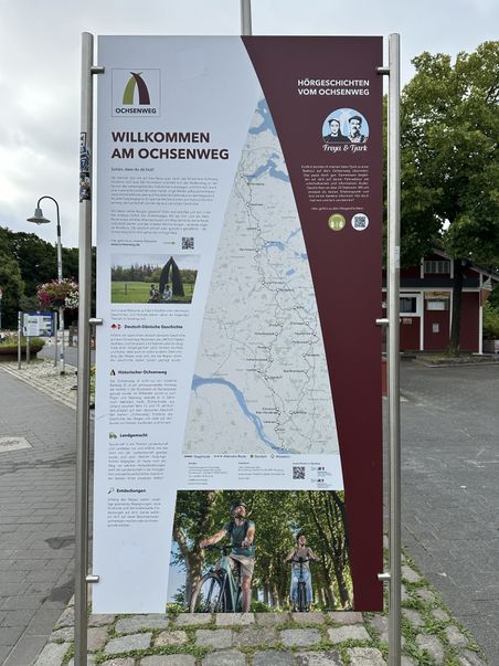 Ein Informationsschild am Ochsenweg zeigt eine Karte und beschreibt die Route. Es gibt Hinweise auf Hörgeschichten und Sehenswürdigkeiten entlang des Weges.