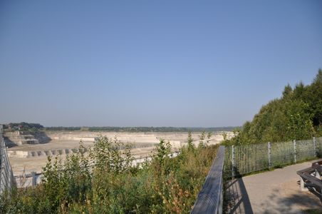Ein Aussichtspunkt mit Blick auf einen großen Steinbruch. Im Vordergrund sind Pflanzen und ein Zaun zu sehen, rechts ein Weg mit Sitzbank.