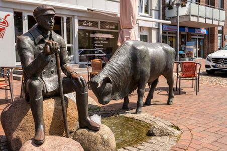 Bronzestatue eines Mannes mit Stock neben einem Stier auf einem Platz in Hohenwestedt. Im Hintergrund ein Café und ein Reisebüro.