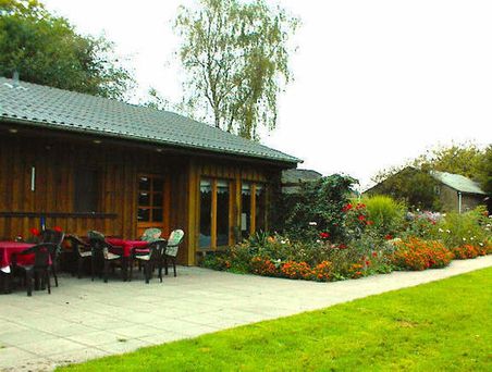 Außenansicht eines Holzgebäudes mit Terrasse, Tischen und Stühlen, Blumenbeet und Rasen.