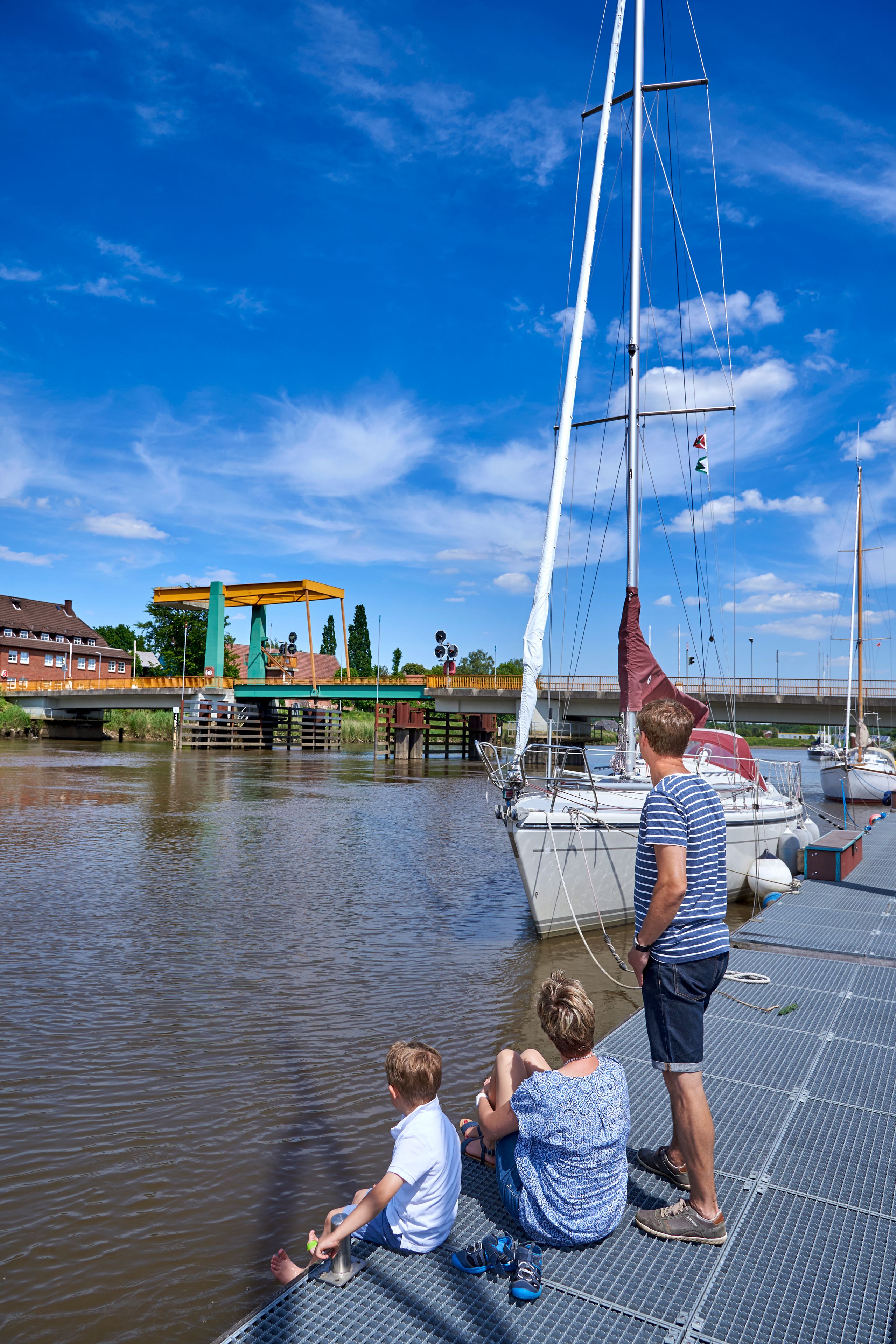 Eine Familie genießt den sonnigen Tag am Hafen von Heiligenstedten. Ein Segelboot liegt am Steg, während die Klappbrücke im Hintergrund zu sehen ist.
