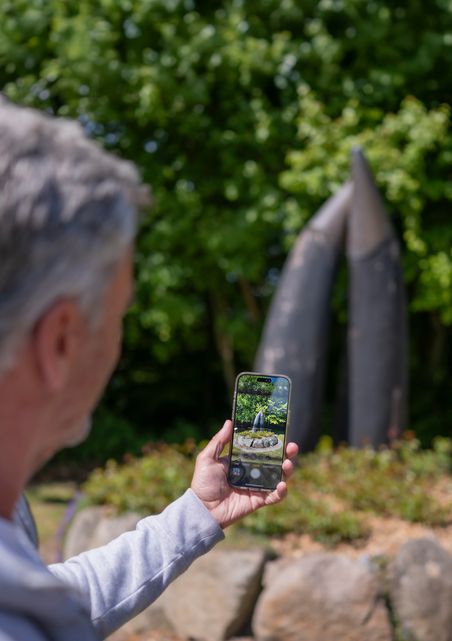 Ein Mann hält ein Smartphone und fotografiert die Hörner im Freien. Die Skulptur besteht aus zwei spitzen, metallischen Formen vor grünem Hintergrund.