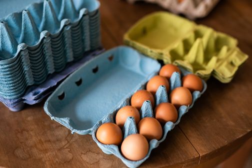 Frische Eier in blauen Kartons auf einem Holztisch. Im Hintergrund sind weitere leere Kartons in verschiedenen Farben gestapelt.