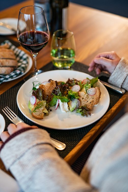 Ein Teller mit frischem Salat, Radieschen und Brotstücken, begleitet von einem Glas Rotwein auf einem gedeckten Tisch.
